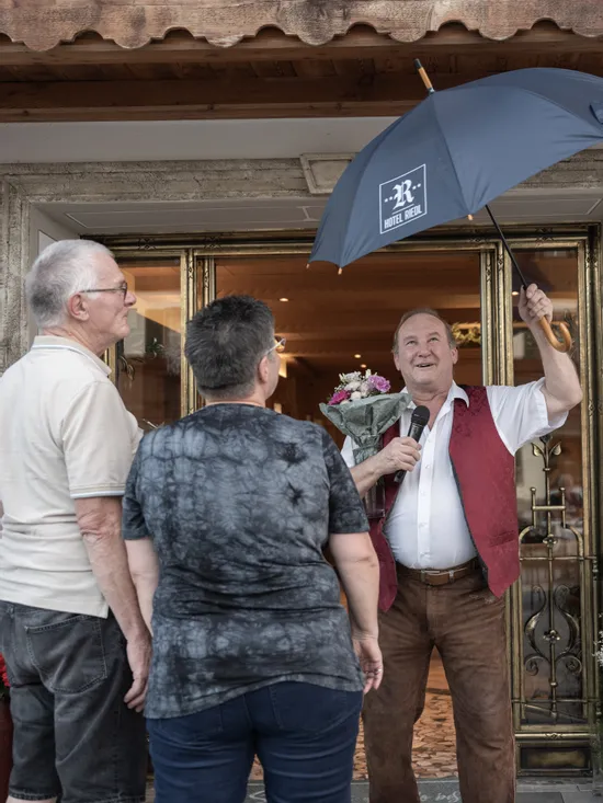 Mann mit Hotel Riedl Regenschirm und Blumenstrauß begrüßt zwei Gäste am Eingang